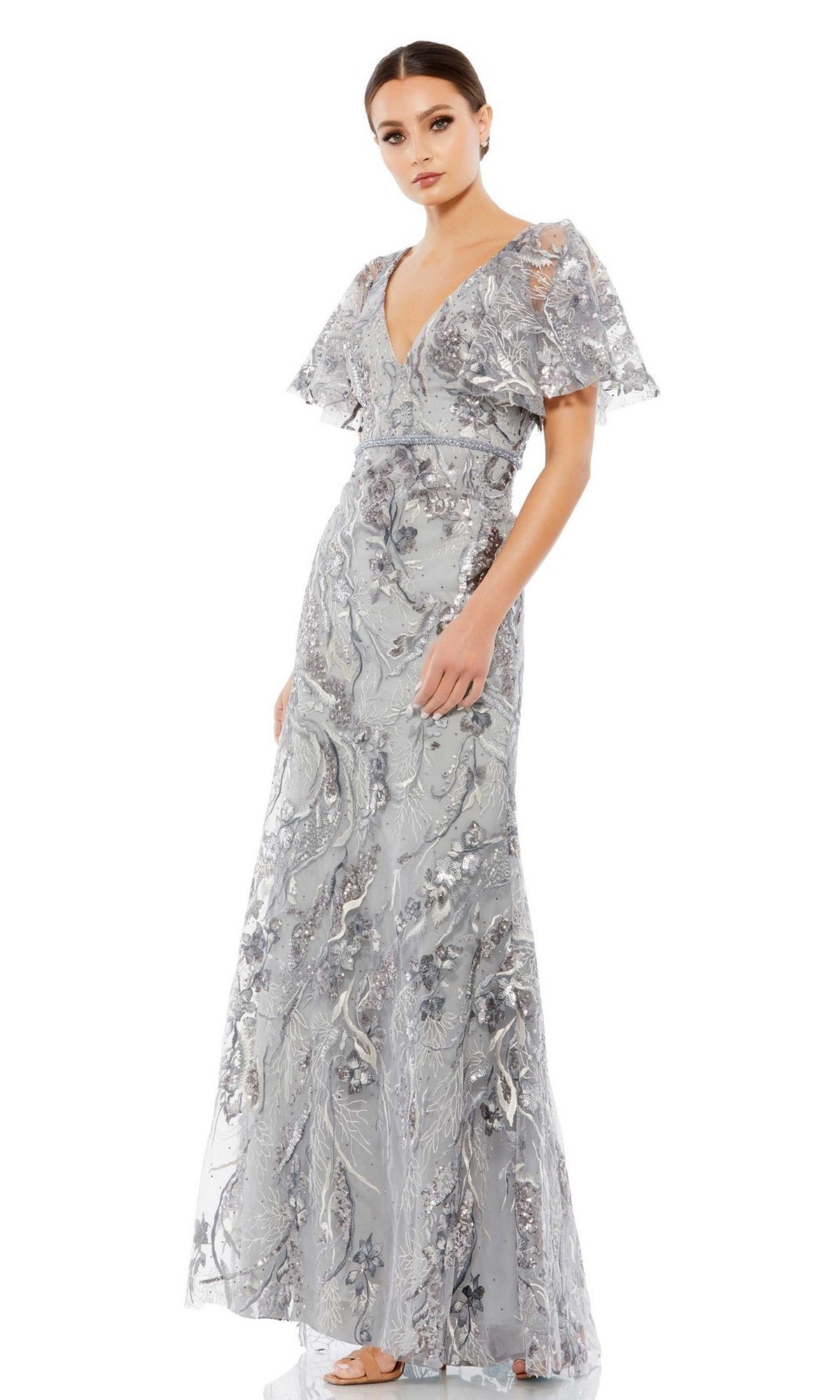 A woman models the Long Formal Dress 67908 by Mac Duggal, featuring silver-grey embroidery with floral patterns and flutter sleeves, standing against a white background.